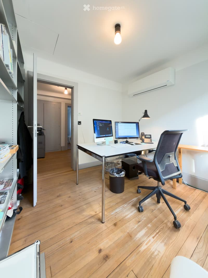 Modern office space with wooden flooring, a white desk, ergonomic chair, dual monitors, and a wall-mounted lamp.