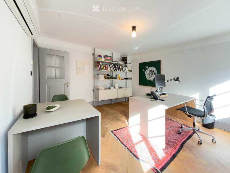 Modern office space with wooden flooring, white walls, and large windows. Features a desk with a computer, two chairs, a bookshelf, and decorative items.
