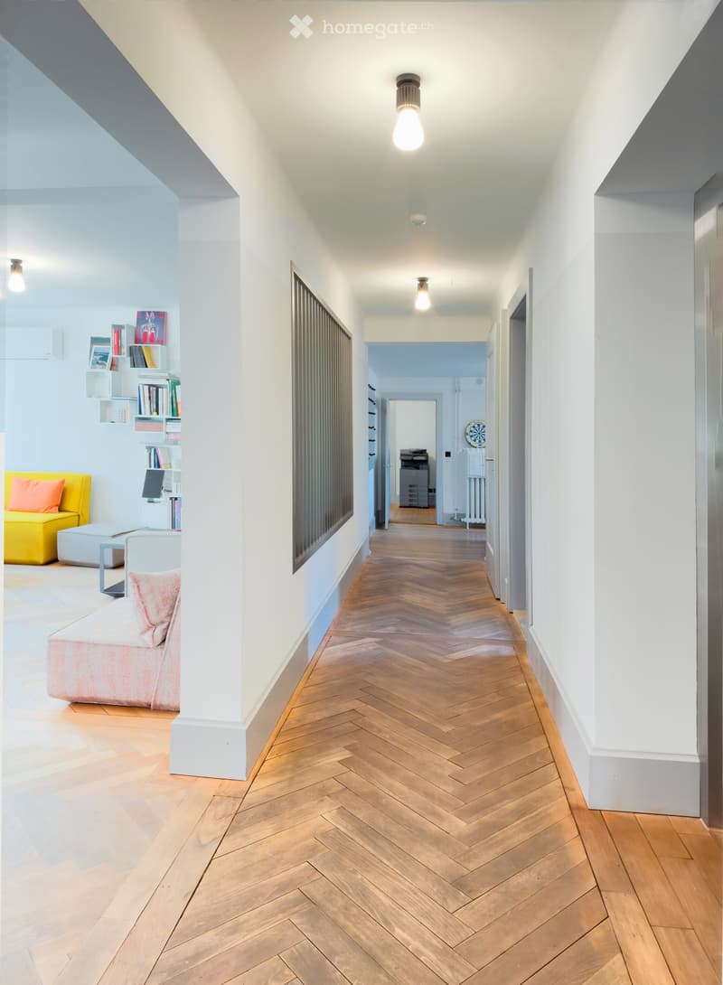 Modern hallway with herringbone wooden flooring, white walls, and pendant lights. Visible rooms include a living area with a yellow sofa and a study area with shelves and a desk.