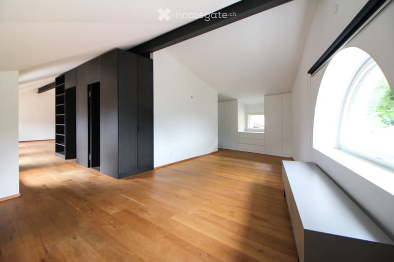 An empty room with wooden flooring, white walls, and large windows allowing natural light. Features include built-in dark grey cabinets and a white bench along the window wall.