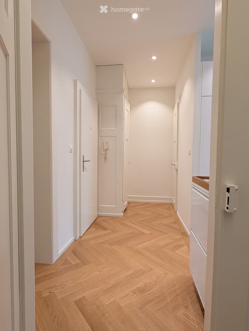 A narrow hallway with wooden herringbone flooring, white walls, and recessed ceiling lights. Doors lead to adjacent rooms, and a small section of a kitchen is visible on the right.