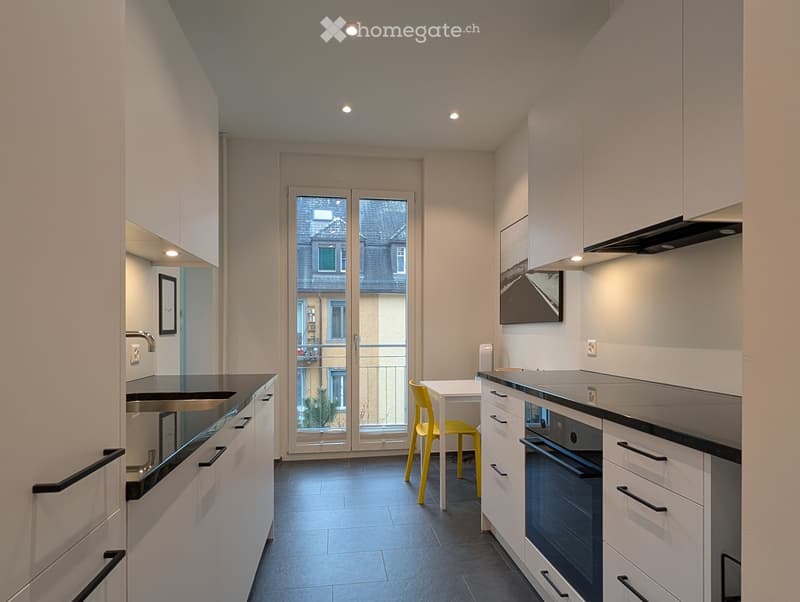 Modern kitchen with white cabinets, black countertops, stainless steel sink, oven, and a small dining area with a yellow chair and white table. A large window offers a view of the street.