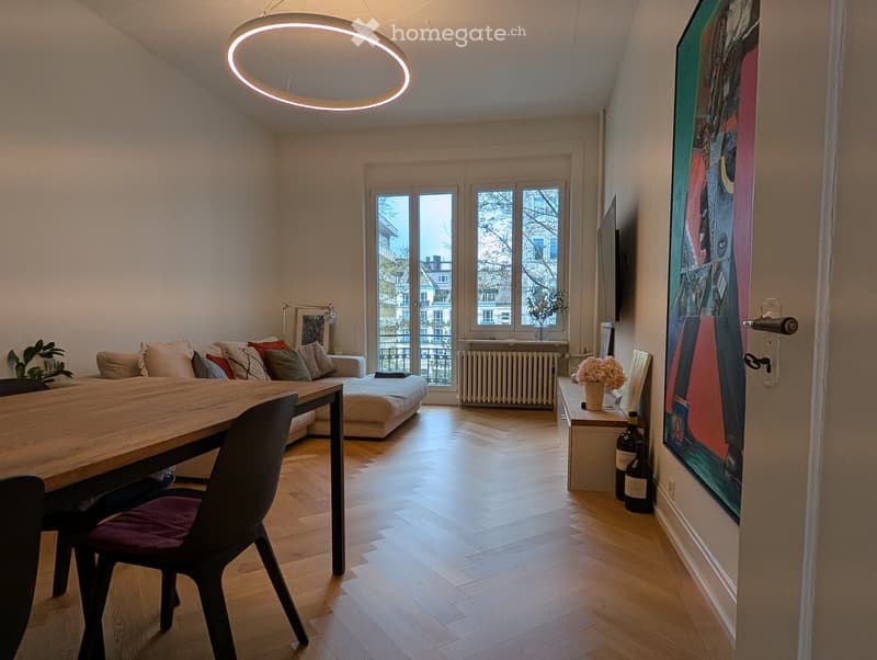 Modern living room with a sofa, dining table, chair, potted plant, artwork, and a bottle on the floor.