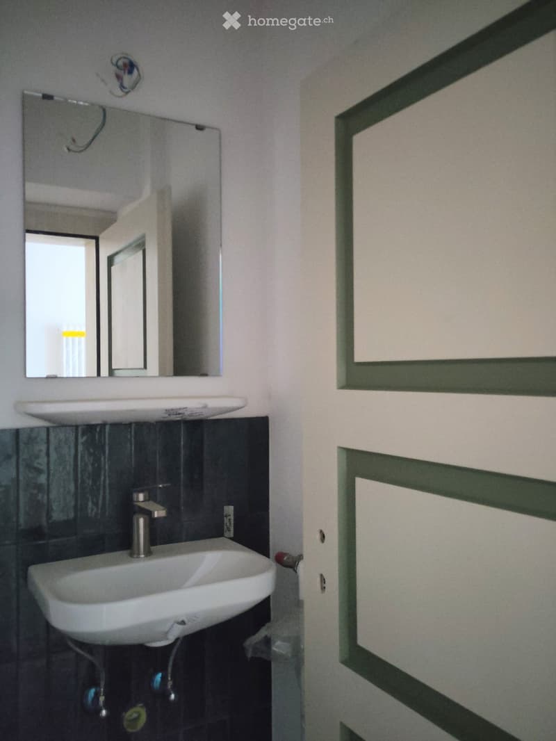 Bathroom with a white sink, chrome faucet, and a rectangular mirror above it. Dark tiled wall behind the sink.
