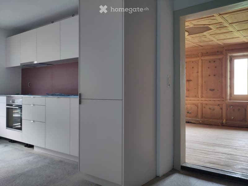Modern kitchen with white cabinets, stainless steel oven, and a doorway leading to a wooden-paneled room.