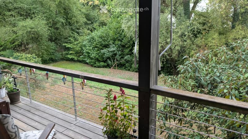 Balcony with wooden decking, metal railing, potted plants, and colorful hanging ornaments. Offers a view of a lush green garden.
