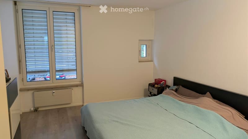 Cozy bedroom with a double bed, nightstand, radiator, and large window with blinds.