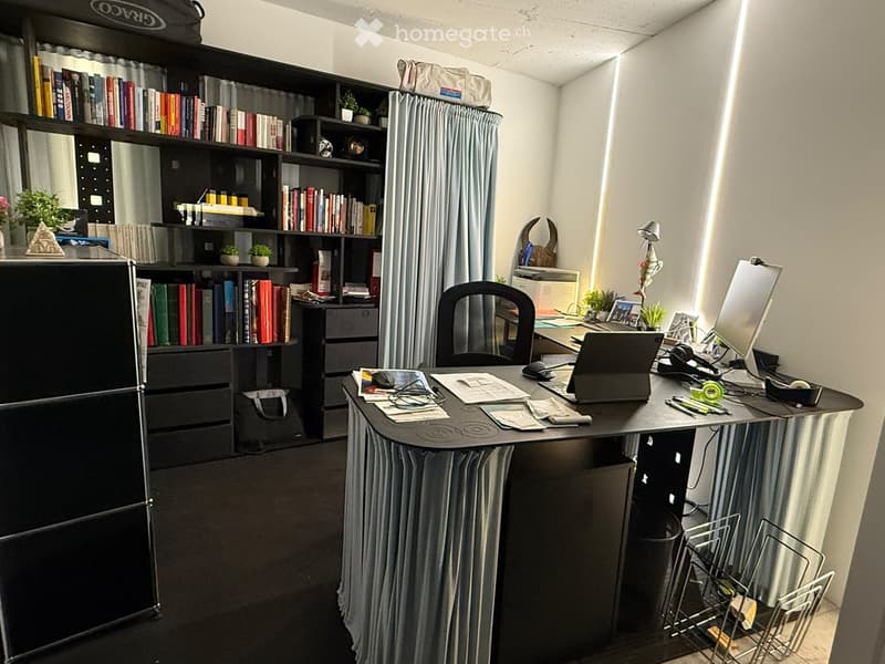 A well-organized home office with a large desk, laptop, screen, bookshelf filled with books, and various office supplies.