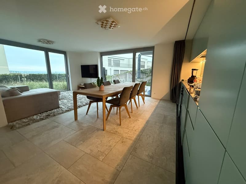 Modern open-plan living and dining area with large windows offering a scenic view, featuring a sofa, dining table with chairs, television, and a kitchen area with cabinets and appliances.