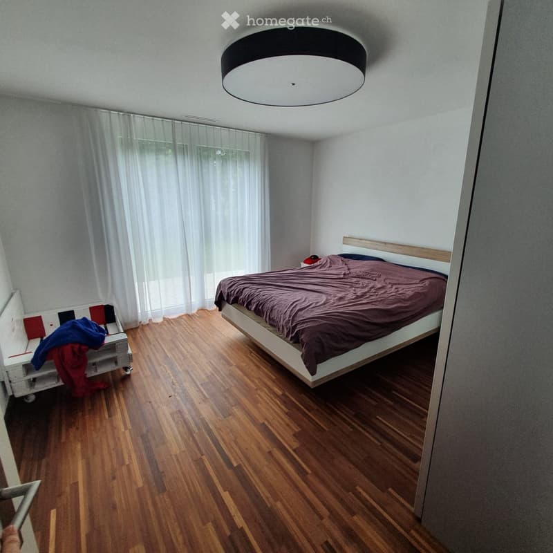 Modern bedroom with wooden flooring, a large bed with dark bedding, a nightstand with a lamp, and sheer white curtains covering large windows.