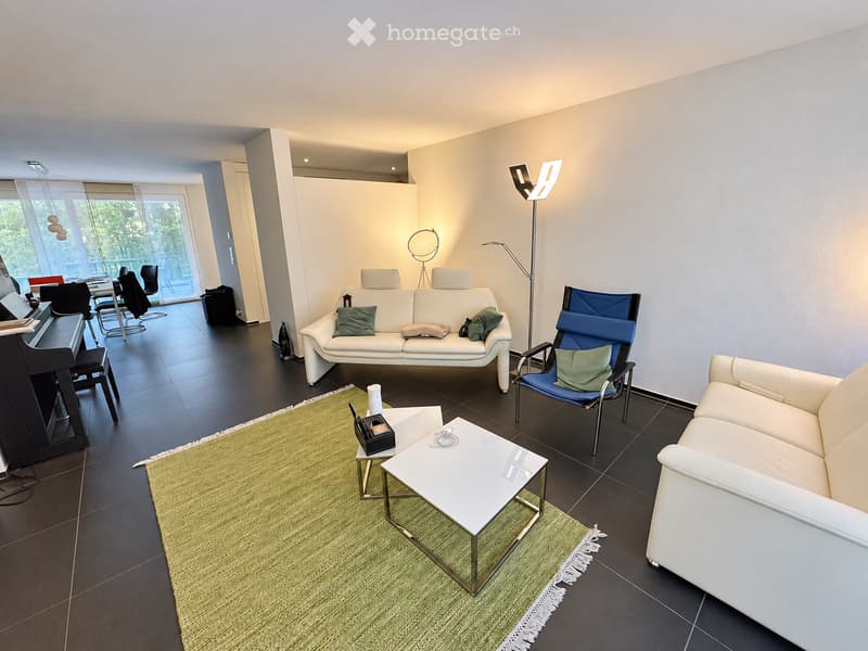 Modern living room with white sofas, a blue accent chair, a white coffee table, a green rug, and a black piano in the corner. Large windows provide natural light.