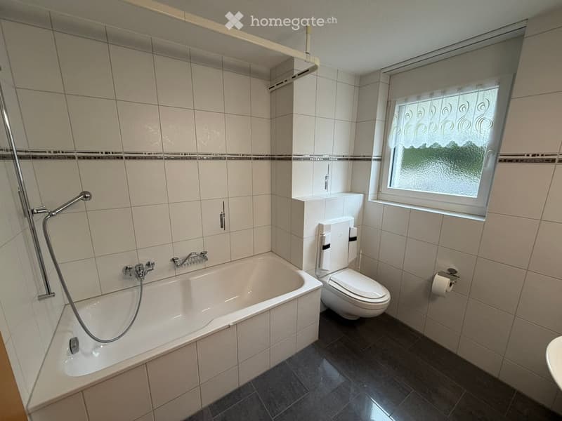 Modern bathroom with a bathtub, toilet, shower hose, and a window with frosted glass. The walls are tiled in light beige with a decorative border, and the floor is dark grey tiles.
