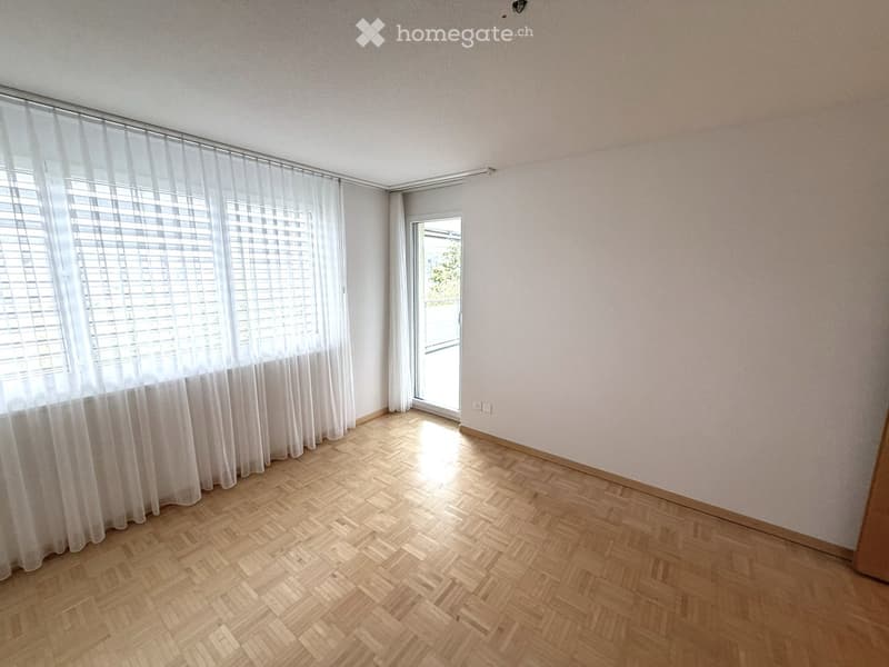 Empty room with wooden herringbone flooring, white walls, large windows with white blinds, and a sliding glass door leading outside.