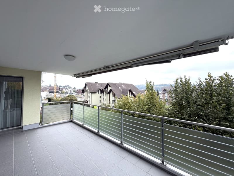 Balcony with modern glass railing, tiled floor, and a retractable awning, overlooking a residential area with trees and buildings.