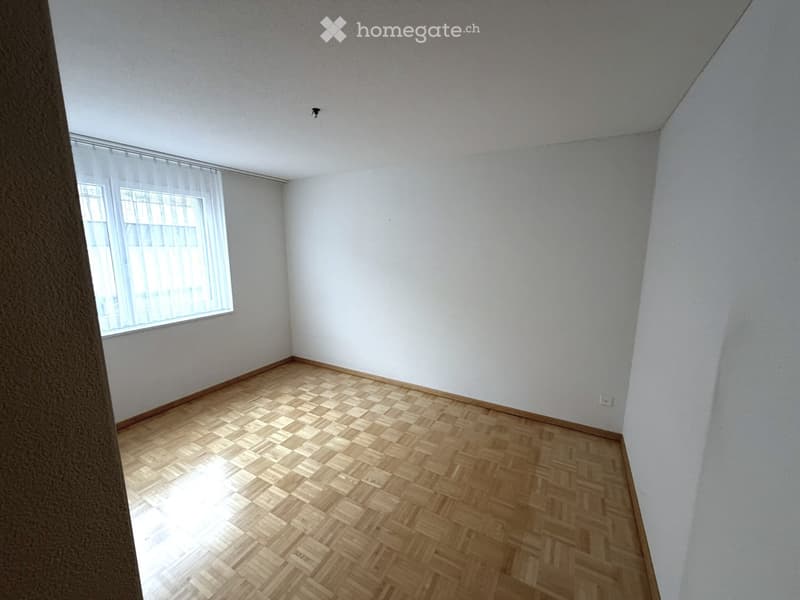 Empty room with wooden parquet flooring, white walls, and a large window with white blinds.