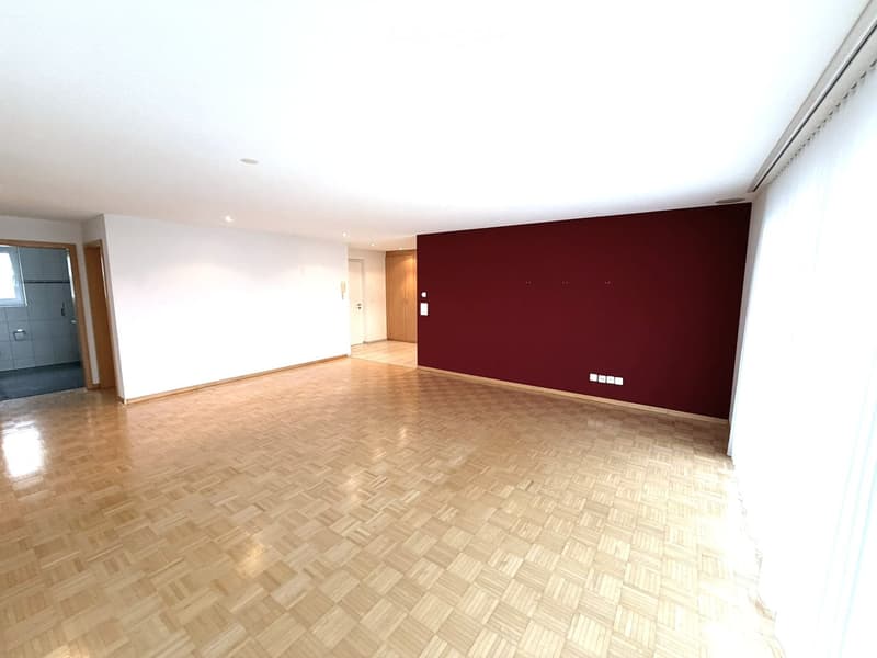 Empty room with wooden herringbone flooring, white walls, and a dark red accent wall. Features include a doorway to another room and a partially visible bathroom with tiled walls.