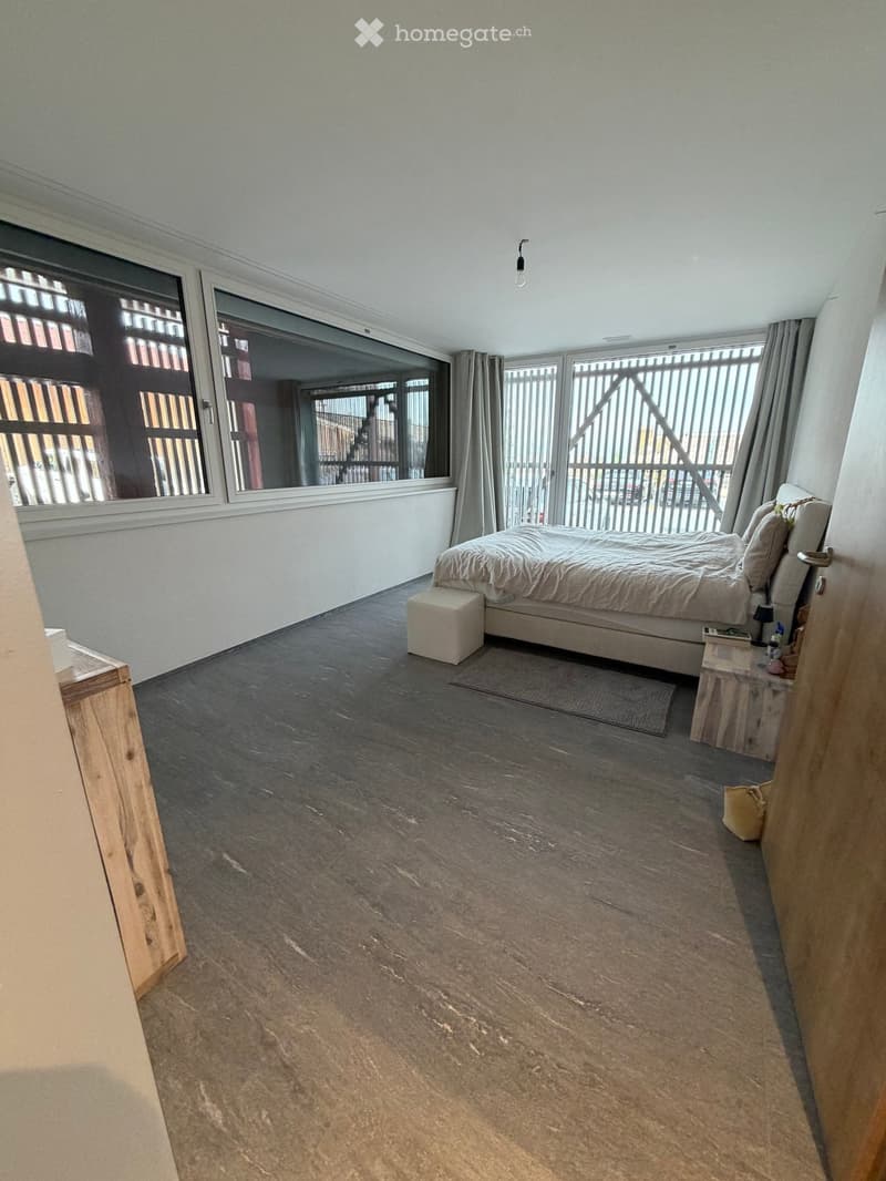 Modern bedroom with large windows, dark wooden flooring, a bed with white bedding, a nightstand, and a small stool.