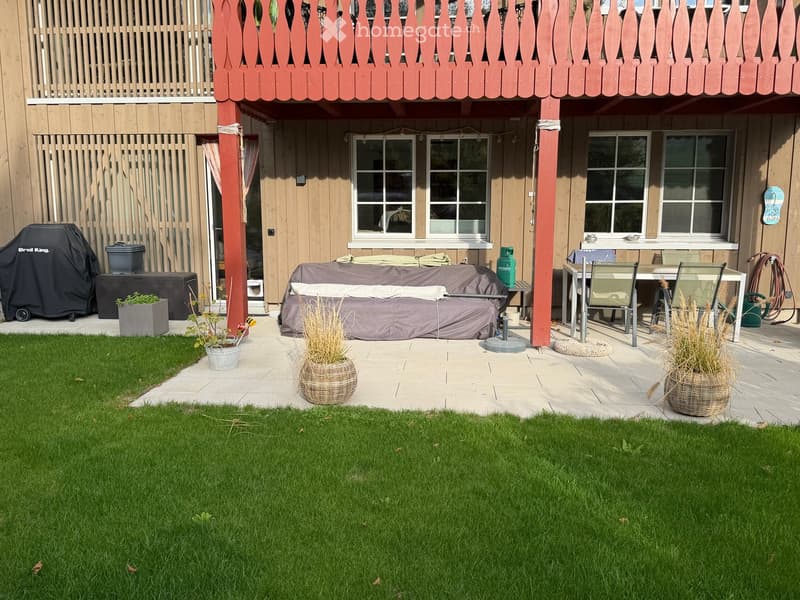 A well-maintained garden with a covered patio area, featuring a table, chairs, a covered sofa, a grill, and potted plants.