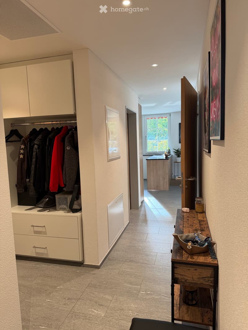 Modern hallway with tiled flooring, built-in closet with hanging clothes, small table with decorative items, and a view into an adjacent room with a wooden counter and plants.