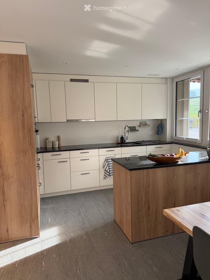 Modern kitchen with white cabinets, black countertops, and a wooden island. Features a sink, faucet, bowl with fruit, and a small dining area with a table and chair.