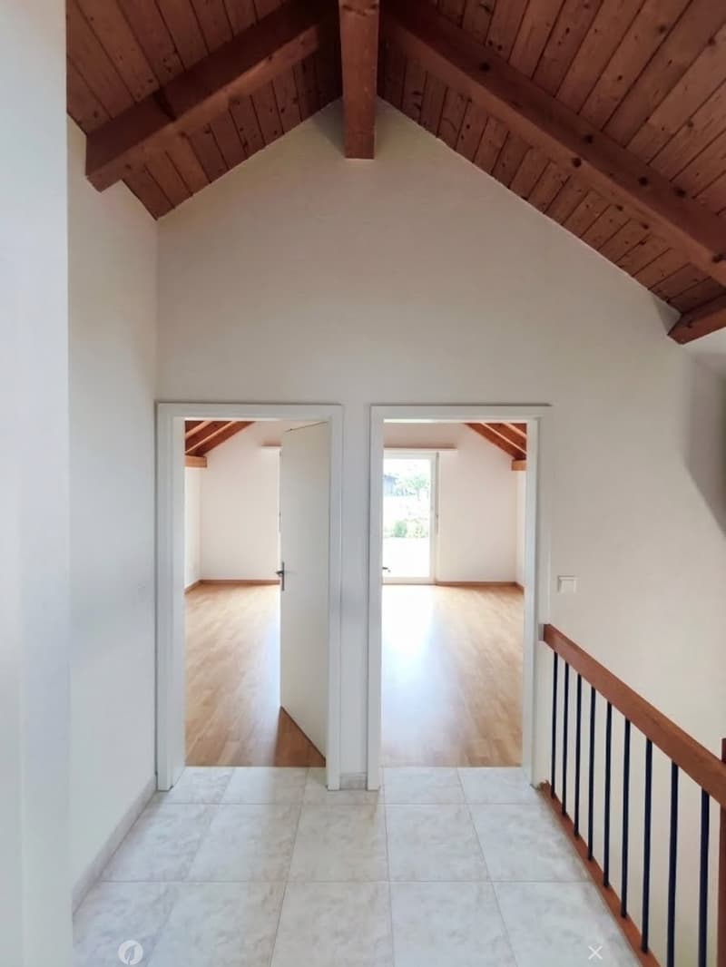 Bright hallway with wooden beams on the ceiling, leading to two open doors that reveal rooms with wooden flooring and white walls.