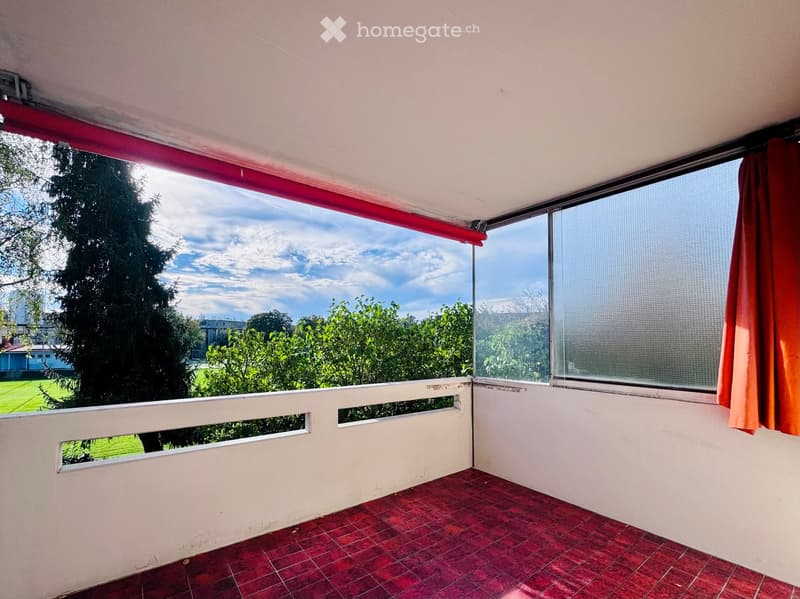 Balcony with red tiled floor, white railing, large windows with blinds, and a view of greenery and a clear sky.