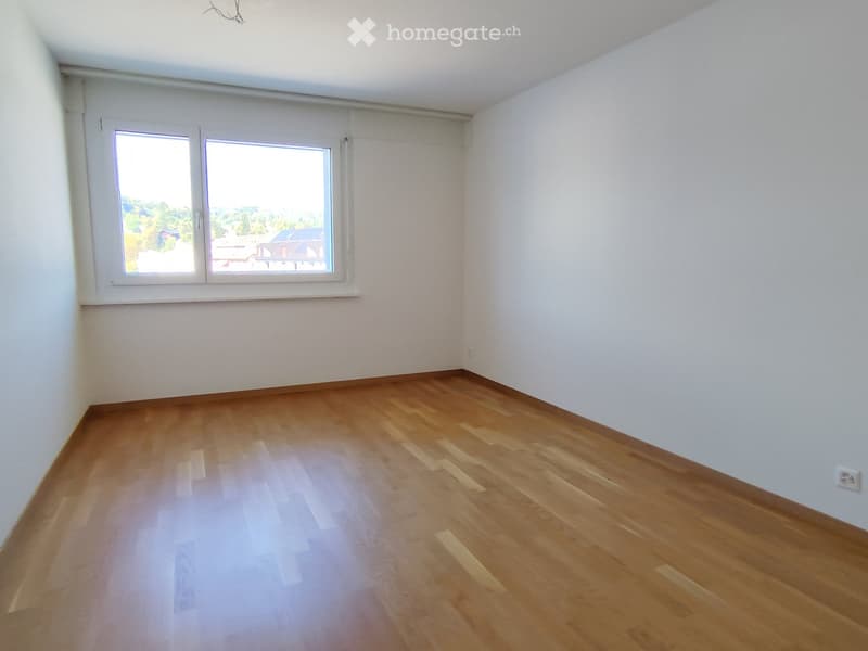 Empty room with wooden flooring and white walls, featuring a large window with a view of greenery outside.