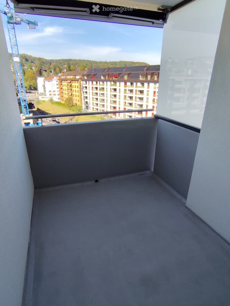 Empty balcony with grey flooring, metal railing, and a view of a cityscape with modern apartment buildings and a construction crane.