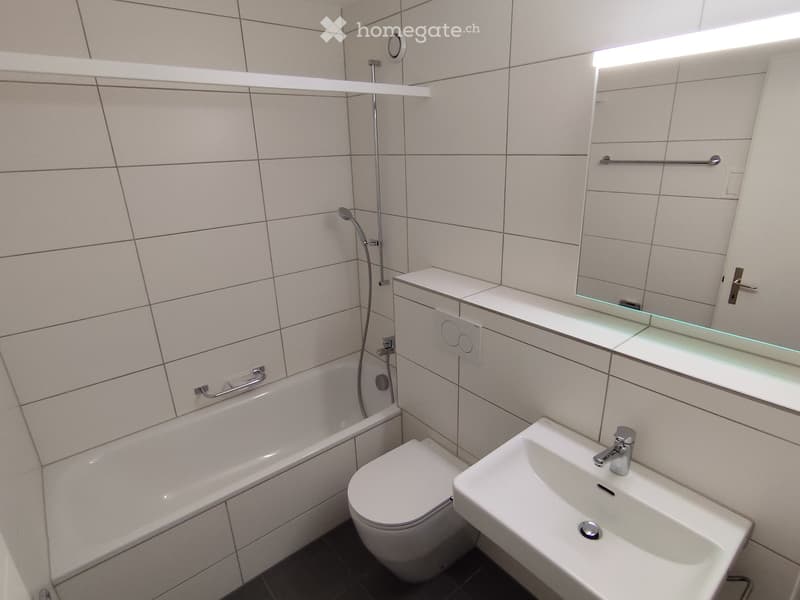 Modern bathroom with a bathtub, toilet, sink, and a glass-fronted mirror with built-in lighting. The walls are tiled in light beige, and the floor is dark.