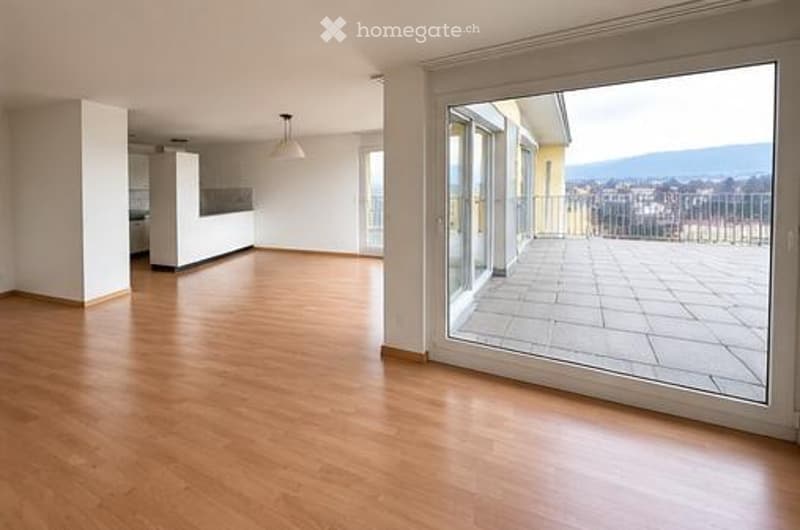 Bright, unfurnished living space with wooden flooring, large sliding glass doors leading to a tiled balcony, and a view of the city and mountains.