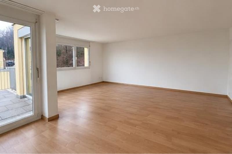 Empty room with wooden flooring, white walls, large windows, and a sliding glass door leading to a balcony.