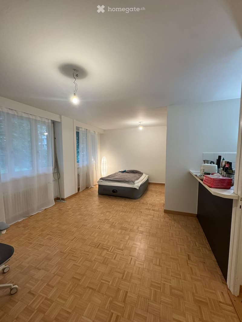 A small, furnished room with a wooden floor, a bed, a chair, a small table with various items, and a lamp near the window.