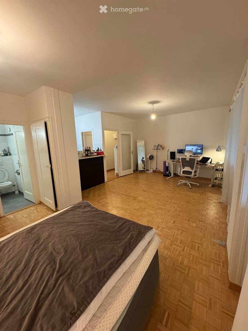 A furnished room with a bed, desk, chair, and a partially visible bathroom. The room features wooden flooring and white walls.