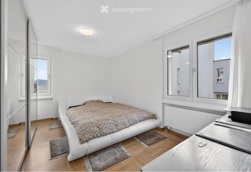 Modern bedroom with white walls, wooden flooring, large windows, a white leather bed with a patterned duvet, a nightstand, and a mirror.