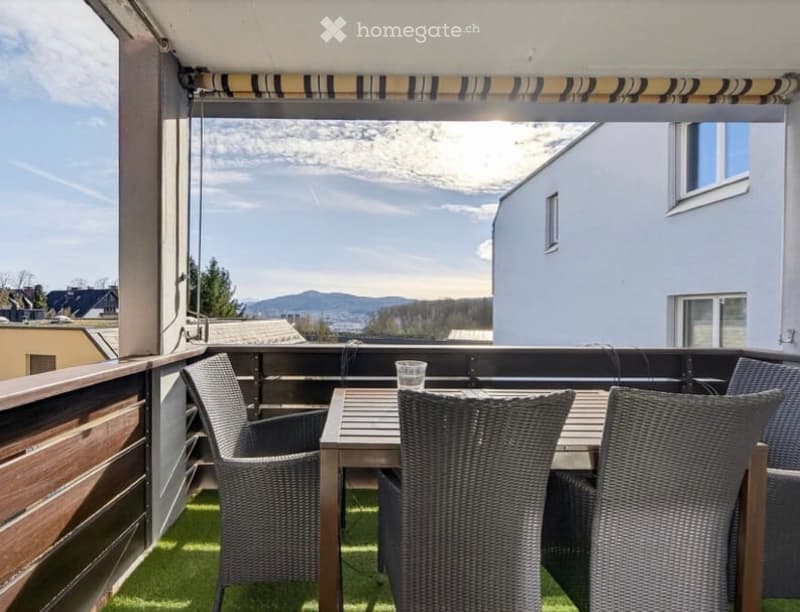 Balcony with a wooden table, wicker chairs, and a glass of water, overlooking a scenic mountain view.