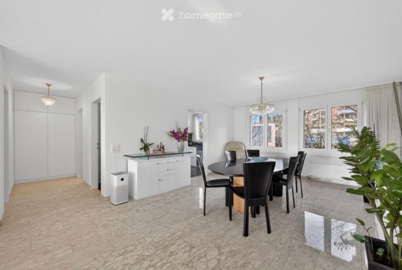 Bright, open dining area with a round glass dining table surrounded by black chairs, a white cabinet with a vase of flowers, and a chandelier hanging from the ceiling. The space features large windows with white curtains and a polished stone floor.
