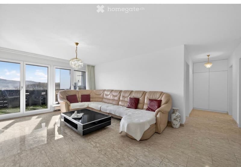 Bright living room with large windows offering a scenic view, featuring a cream-colored sectional sofa with red throw pillows, a black coffee table, and a decorative vase near the window.