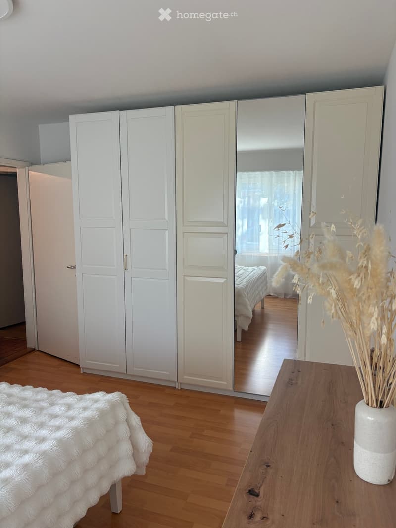 Modern bedroom with wooden flooring, large white wardrobe with mirrored doors, a bed with a white fur throw, a wooden side table with a vase of dried plants.