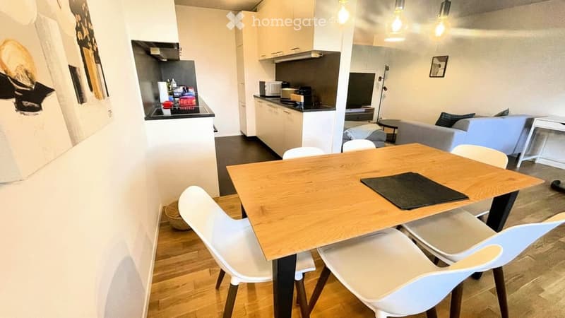Modern, open-plan living and dining area with a wooden dining table, white chairs, a grey sofa, and a small kitchen area with cabinets, a microwave, and a refrigerator.