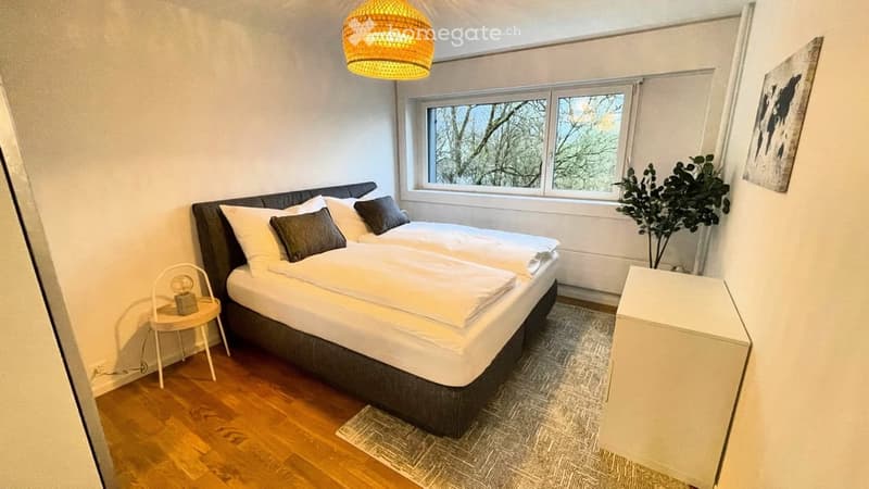 Cozy bedroom with a double bed, nightstand, small round table with a lamp, and a potted plant near the window. The room features wooden flooring and a modern aesthetic.