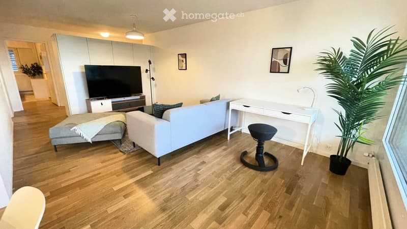 Modern living room with light-colored furniture, including a white sofa, grey ottoman, white desk, black stool, and a large TV stand. The room features wooden flooring, large windows, and decorative elements like a plant and picture frames.