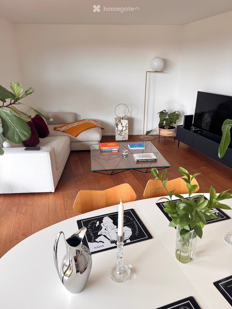 Modern living room with a white sofa, glass coffee table, wooden chairs, and decorative items including candles, a vase, and potted plants.