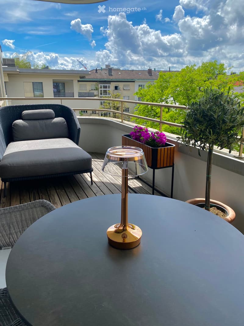 Balcony with wooden decking, grey sofa, round table, wicker chair, flower pot with purple flowers, and a decorative lamp.