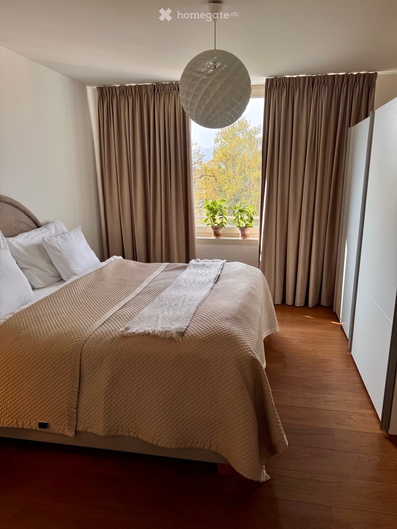 A well-lit bedroom with a large bed, pillows, a blanket, and a decorative throw. The room features wooden flooring, large windows with curtains, and a pendant light hanging from the ceiling. Two potted plants are placed on the windowsill.