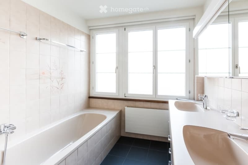 Bright bathroom with a bathtub, double sink, large windows, and tiled walls.