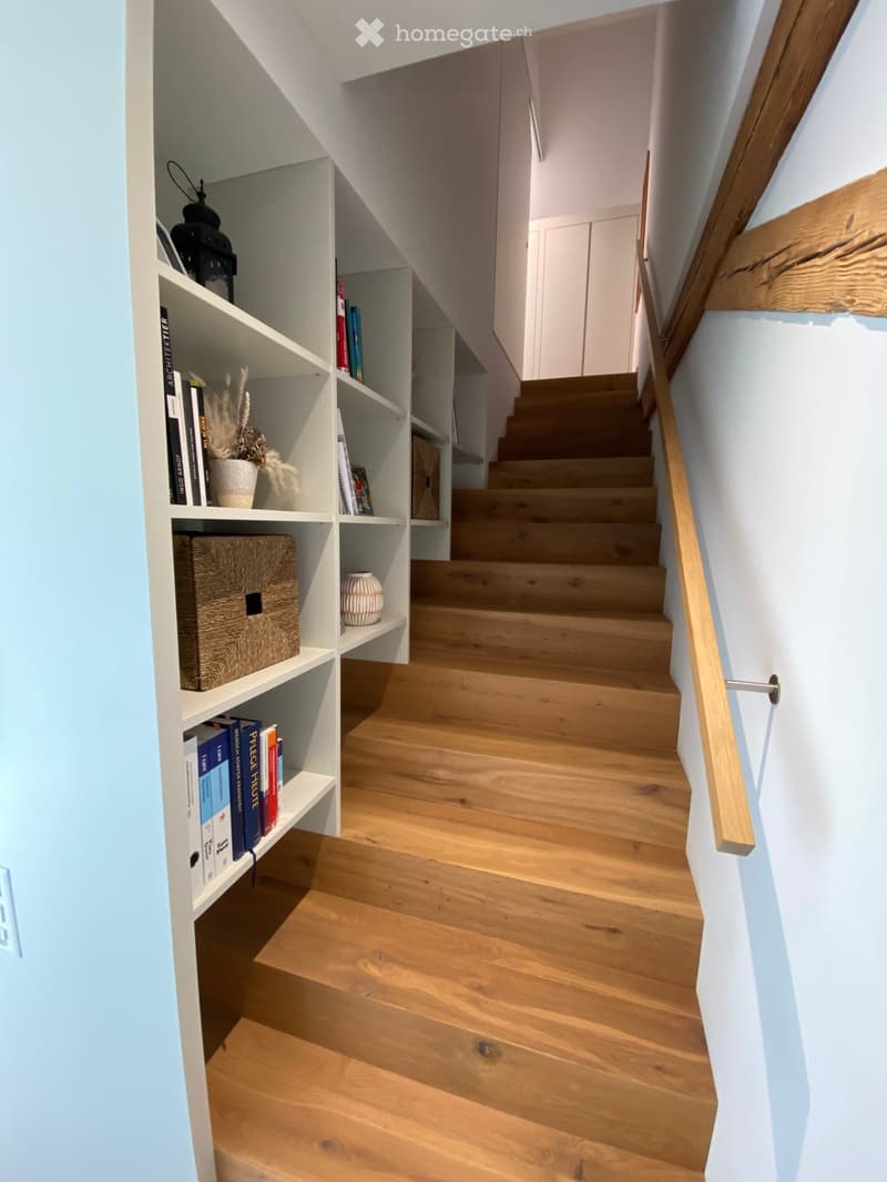Wooden staircase with built-in white shelving unit on one side, containing books, decorative items, and a small plant. The staircase features a wooden handrail and light-colored wooden steps.
