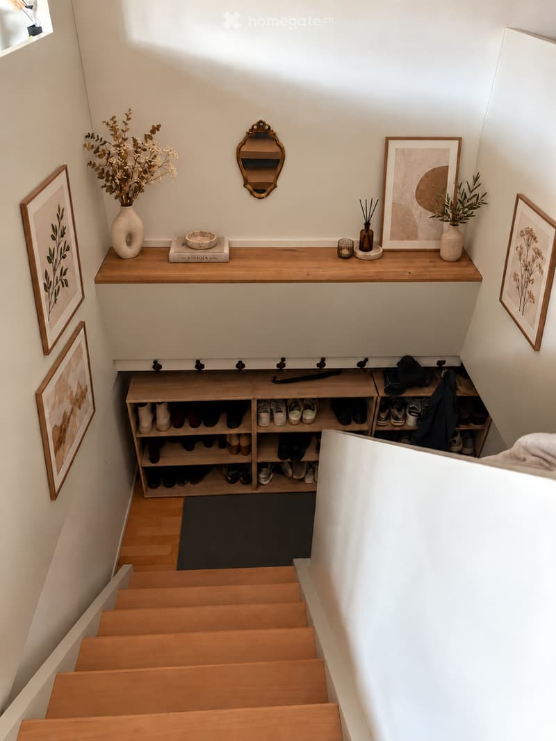 Staircase with wooden steps and a built-in shoe storage unit below. The landing features a wooden shelf with decorative items, including vases, a mirror, and framed artwork.