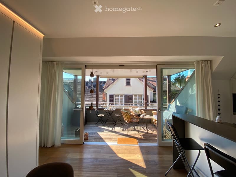 Bright living space with wooden flooring, large glass doors opening to a balcony, sofa, chairs, small tables, and a bar counter.