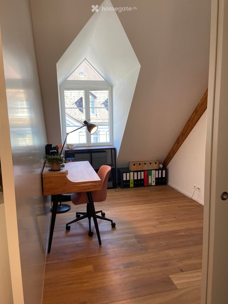Bright attic office with wooden flooring, large skylight window, modern desk, ergonomic chair, and storage units.
