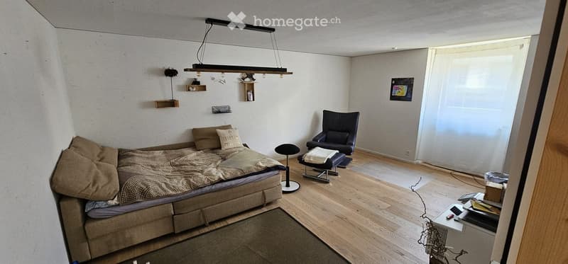 Cozy bedroom with a bed, armchair, small table, shelves, and a hanging light fixture. The room features wooden flooring and white walls.